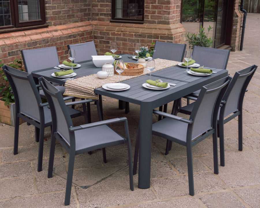 An outdoor dining set with a extending butterfly table and eight stackable chairs, all in light grey color, set on a paved outdoor area with a brick wall in the background.